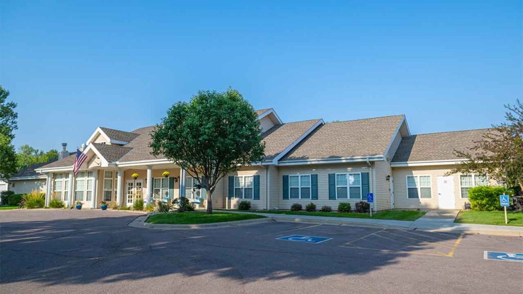Single-story building with light-colored siding, numerous windows, and a central entrance adorned with an American flag. The building is surrounded by a parking lot that includes designated handicap parking spaces and is set against a clear blue sky.