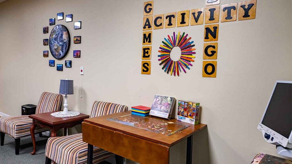 A cozy room with two striped armchairs and a small table with a lamp and doily. Nearby is a wooden table with a jigsaw puzzle, a colorful wheel, and games. The wall above features the words