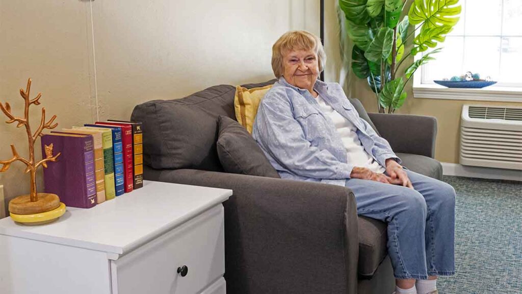 An elderly person with short light hair is sitting on a gray couch in a cozy room. They are smiling and wearing a light blue shirt over a white top and blue pants. A white end table with books and a decorative piece is next to the couch. A large plant is by the window.