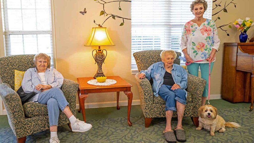Three elderly women are sitting in a living room. Two are seated in armchairs, while the third woman stands, holding a small dog on a leash. The room is decorated with plants, a table lamp, floral wallpaper, and a cabinet with a vase of flowers.