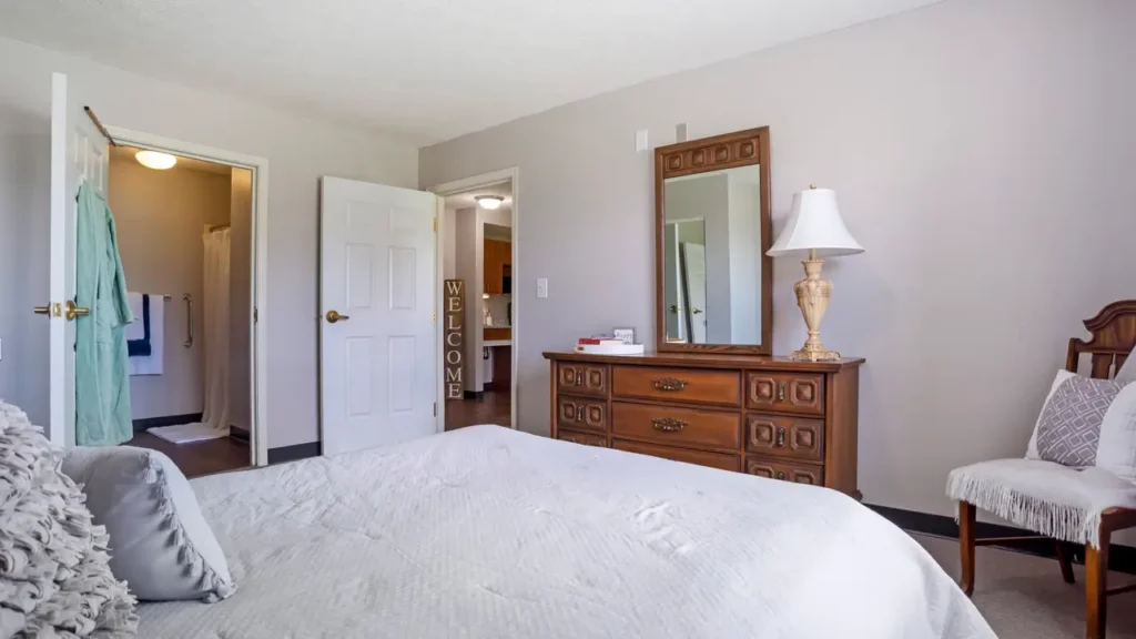 A bright assisted living bedroom with a neatly made bed, wooden dresser with a mirror and lamp, two white chairs, and open doors leading to a bathroom and hallway with a “Welcome” sign. Light walls and natural light fill the room.