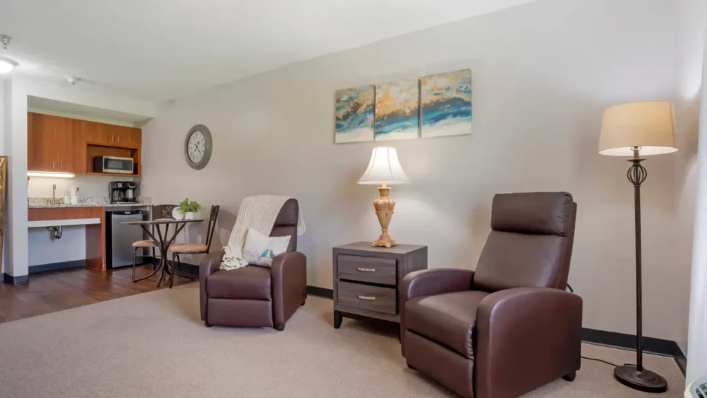 A cozy assisted living space features two brown recliners, a side table with a lamp, a standing floor lamp, wall art, and a clock. In the background, there is a small dining table and a kitchenette area.