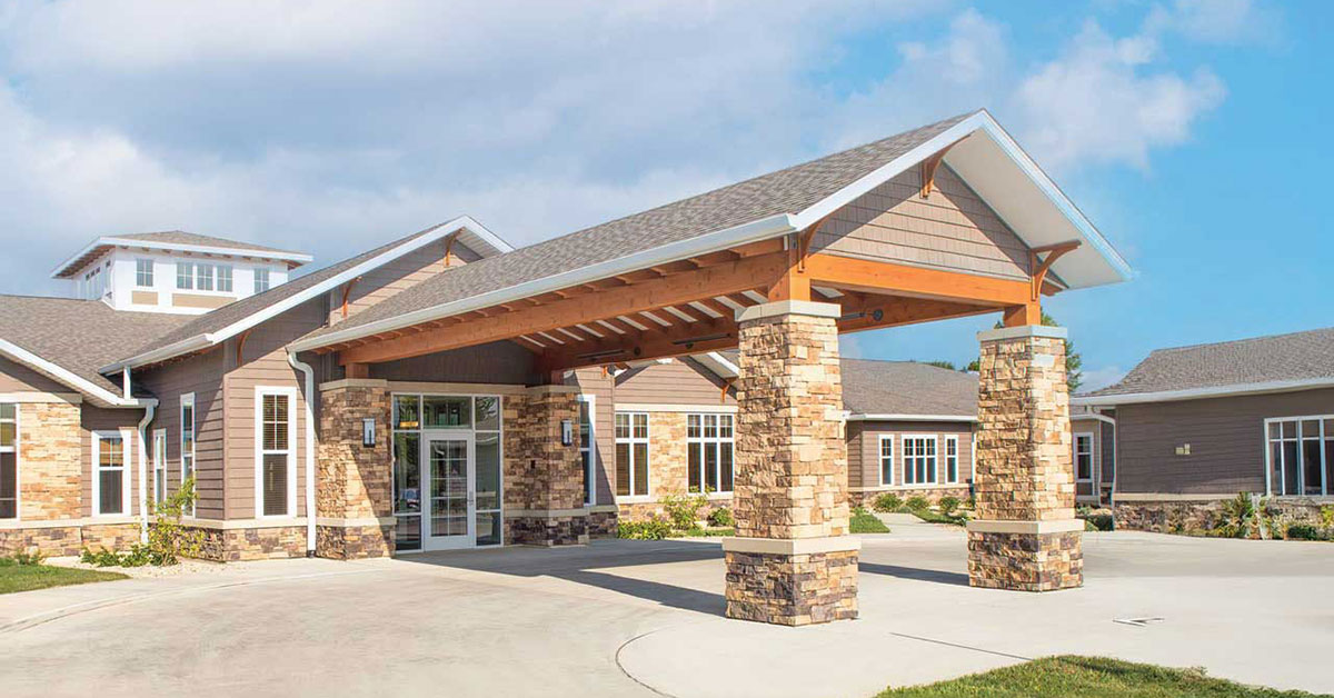 A modern, single-story building with light brown walls and large windows features a covered entrance supported by stone pillars. The sky is clear with scattered clouds, and the surrounding area is landscaped with grass and shrubs.