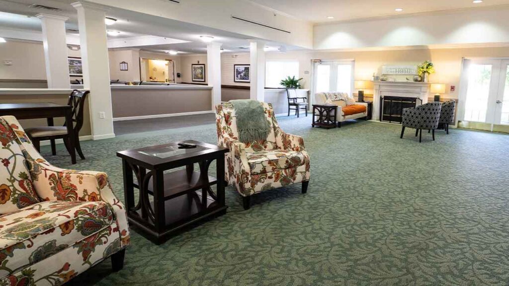 A brightly lit, spacious room with green carpet and white walls. It features floral upholstered chairs, dark wooden side tables, and a cozy seating group around a fireplace. Potted plants and wall art provide homey touches, and large windows let in natural light.