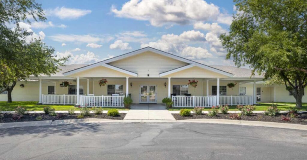 Single-story building with a white exterior and a wide front porch, surrounded by green lawns, trees, and small landscaped bushes, under a partly cloudy sky.