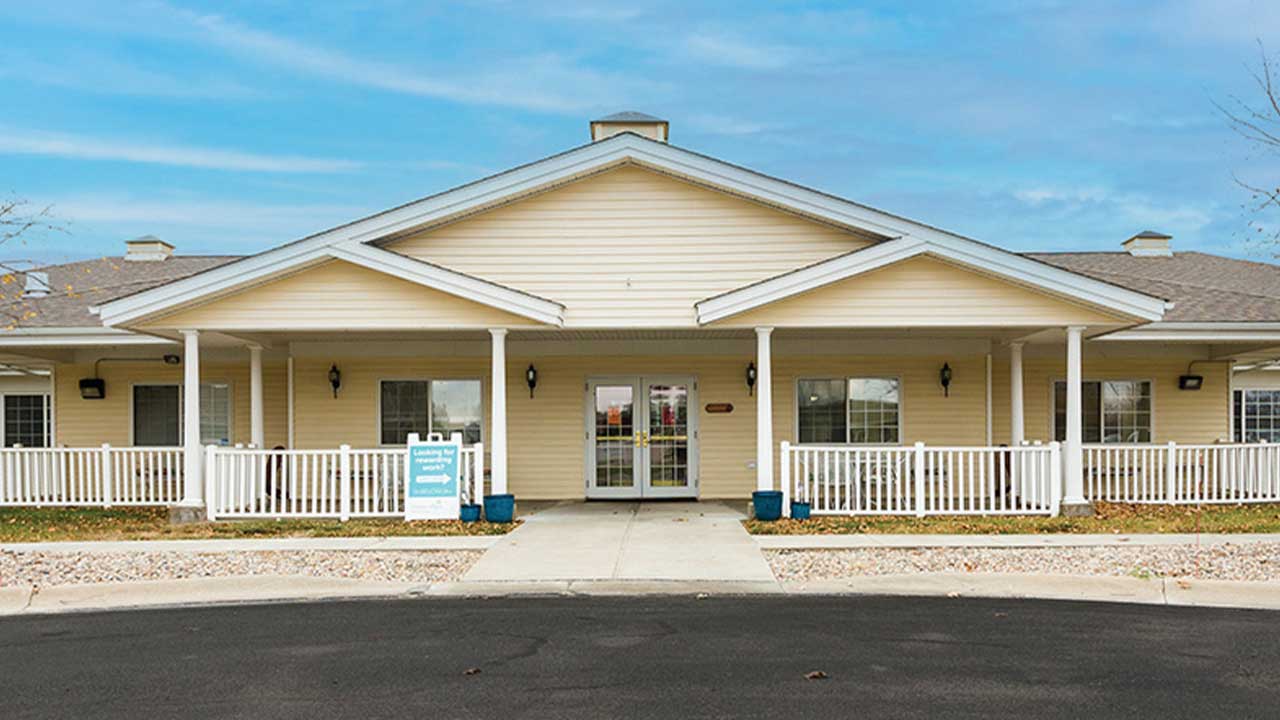 A single-story building with beige siding and a prominent covered entrance featuring white pillars and railings. A signboard is placed near the entrance. The area in front is paved with a walkway leading to the double glass doors and a neat lawn on either side.