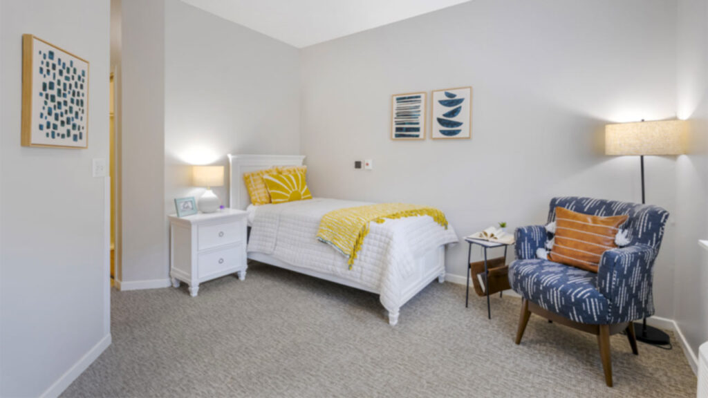 A cozy bedroom with a single bed, white bedding, and yellow accents. A patterned blue armchair with an orange pillow sits beside a lamp. Two abstract pictures hang on the light gray walls.