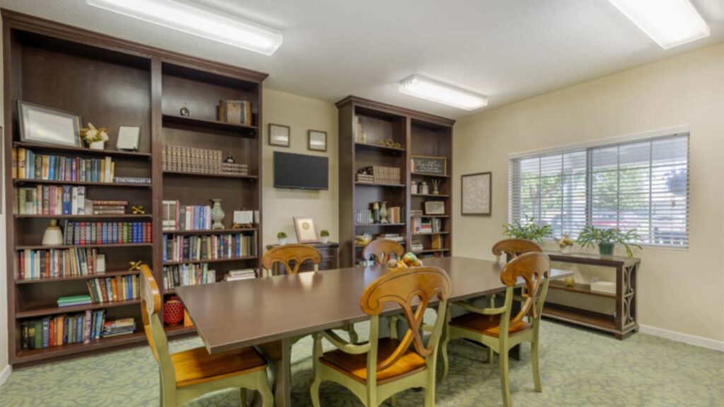 A well-lit library room with a large wooden table surrounded by six chairs, built-in bookshelves filled with books and decor, and a window letting in natural light.