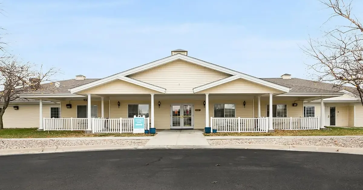 A single-story beige building with a front porch and white railings, featuring a central peaked roof. The entrance is in the middle with double glass doors, flanked by windows. The yard has bare trees and a paved driveway in the foreground.