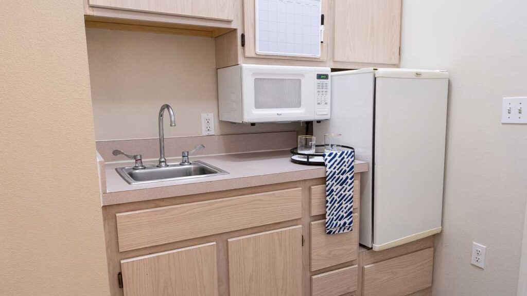 A small kitchen with light wood cabinets, a compact refrigerator, a microwave atop the fridge, a single-basin sink with a faucet, and a blue and white tea towel hanging from the counter. A white notice board is mounted above the microwave.