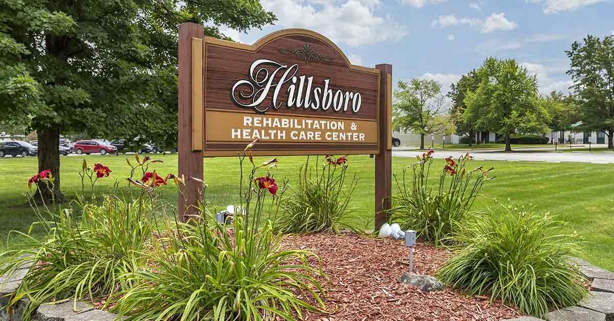 A wooden sign reading "Hillsboro Rehabilitation & Health Care Center" stands in a landscaped area with flowers and greenery, highlighting the inviting grounds of the Rehabilitation & Health Care Center beneath a partly cloudy sky.