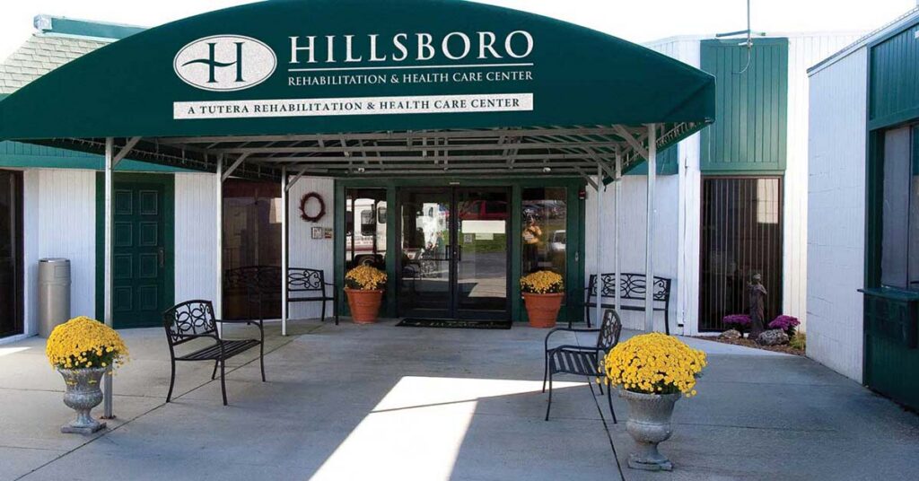 The entrance to Hillsboro Rehabilitation & Health Care Center features a green awning, glass double doors, benches, decorative chairs, and yellow potted flowers, welcoming guests to the center on a sunny day.