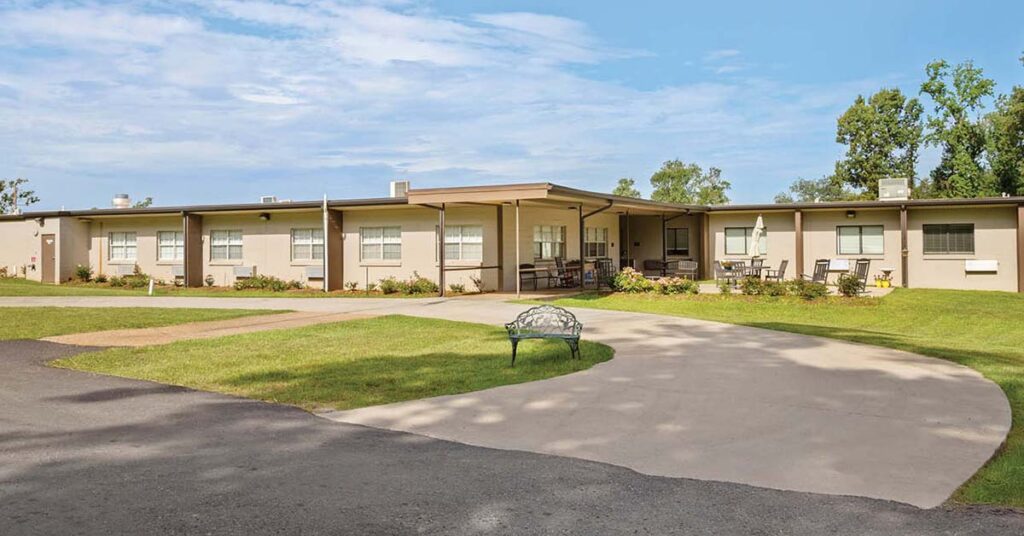Single-story beige Nursing & Rehabilitation building with multiple windows, surrounded by green grass and trees. A curved driveway leads to the entrance, with a decorative metal bench placed on the lawn in front.