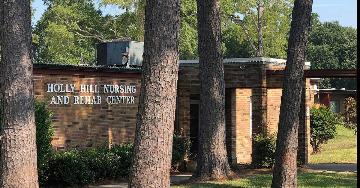 A brick building surrounded by trees features "Holly Hill Nursing & Rehabilitation Center" on its facade. Lush green grass and shrubs fill the foreground, set against a backdrop of a bright blue sky.