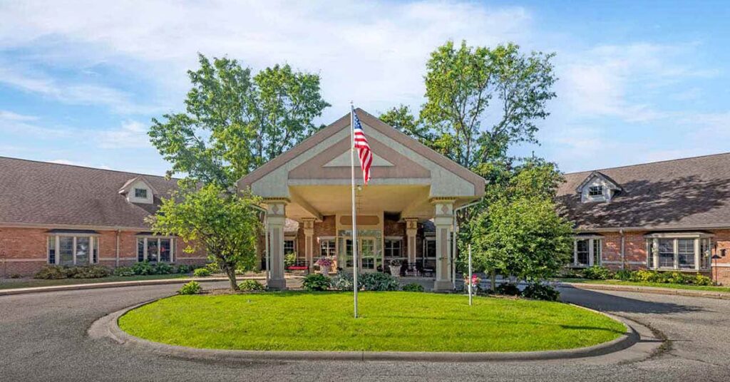 A single-story brick building with a covered entrance, surrounded by trees and bushes, features an American flag in the center of a circular driveway under a blue sky.
