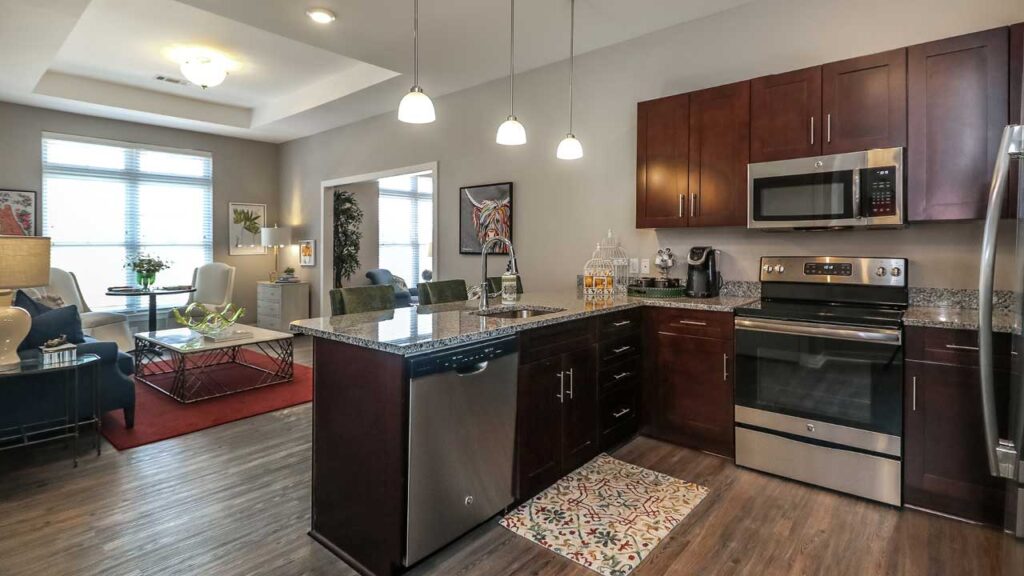 Modern kitchen with dark wood cabinets, granite countertops, and stainless steel appliances. The floor is gray wood, and the kitchen island has a dishwasher with pendant lights above. An open living and dining area is visible in the background, perfectly designed for senior living convenience and style.