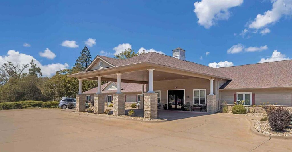 A single-story building with a covered entrance supported by columns, a paved driveway, and landscaped areas with bushes and trees under a blue sky with scattered clouds.