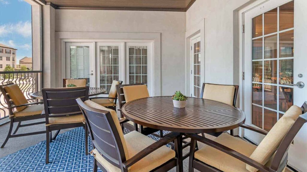 A covered balcony in our Assisted Living & Memory Care community features a round wooden table, six cushioned chairs, a potted plant centerpiece, glass doors, and a blue patterned rug, overlooking nearby buildings on a sunny day.