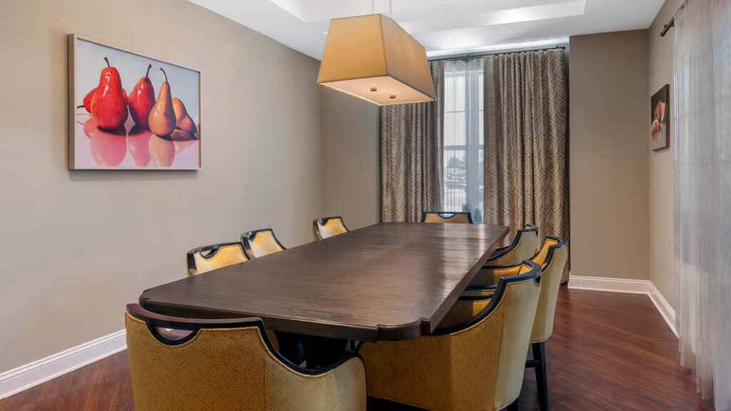 A modern Assisted Living & Memory Care dining room with a dark wooden table, eight yellow upholstered chairs, a large square pendant light, patterned curtains on the window, and framed pear artwork on the wall.