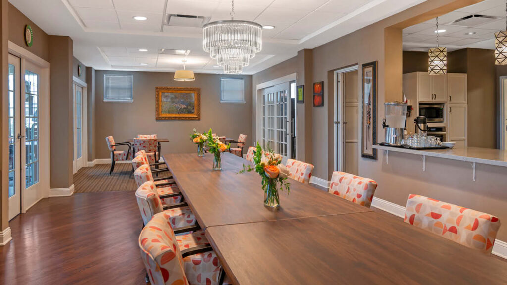 A modern dining room in this Assisted Living & Memory Care community features a long wooden table, patterned chairs, floral centerpieces, and chandelier lighting. Artwork and neutral walls create a warm, inviting atmosphere with a view into the kitchen area.