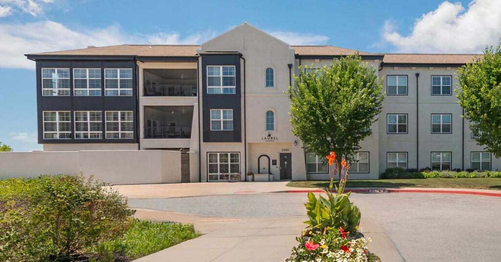 A modern, three-story Assisted Living & Memory Care apartment building with a beige and dark gray exterior, large windows, balconies, and landscaped grounds filled with flowering plants under a partly cloudy sky.