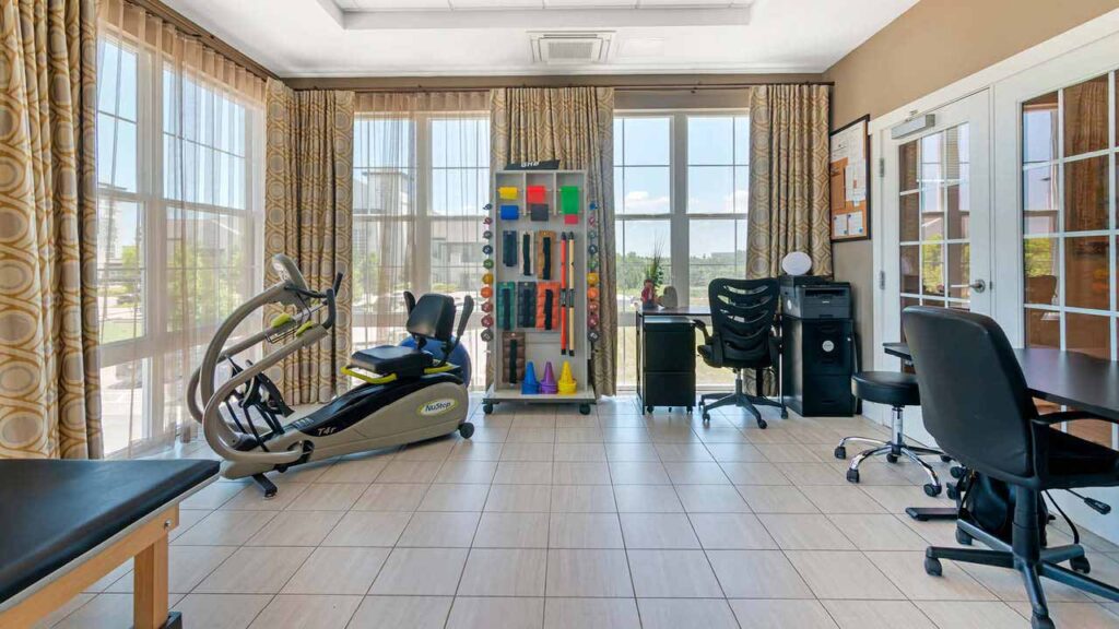 A bright Assisted Living & Memory Care physical therapy room with large windows, exercise equipment, a recumbent bike, therapy tools, colorful cones, desks, chairs, a computer, and a printer—all on a tiled floor with beige curtains.