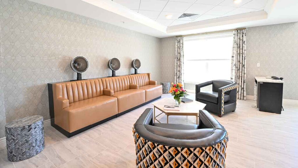 A modern hair salon waiting area featuring brown leather seating with built-in hair dryers, two stylish armchairs with patterned fabric, a small coffee table with a floral arrangement, and large windows letting in natural light. The walls and floor have a neutral-toned design.