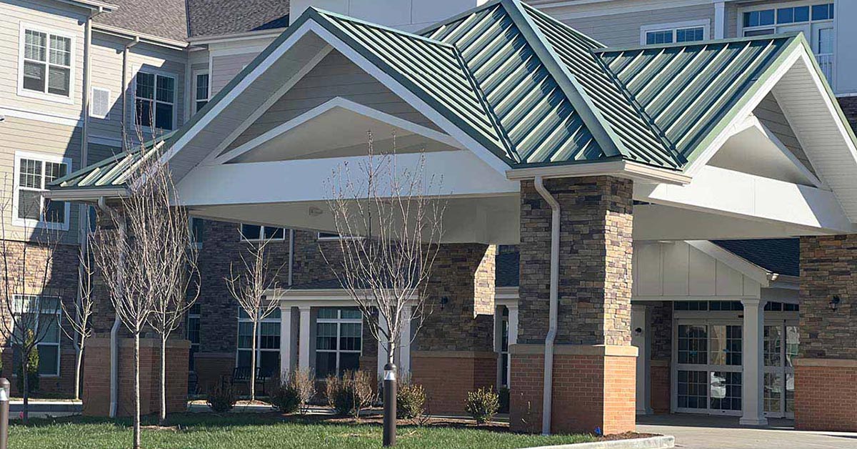 A modern building with a green metal roof and a prominent entrance canopy supported by stone columns. The facade features beige siding and large windows. There are bare trees and grass in the foreground.