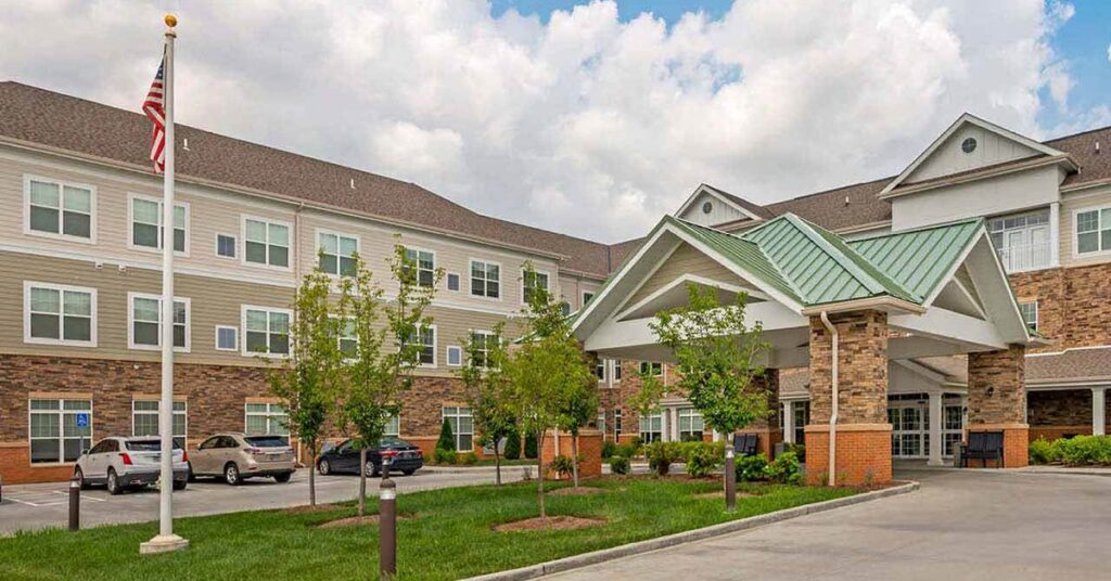 A large, modern, multi-story building with brick and siding offers Senior Living in Chesterfield, MO. It features a covered entrance, several cars parked outside, green trees, an American flag, and a partly cloudy sky.