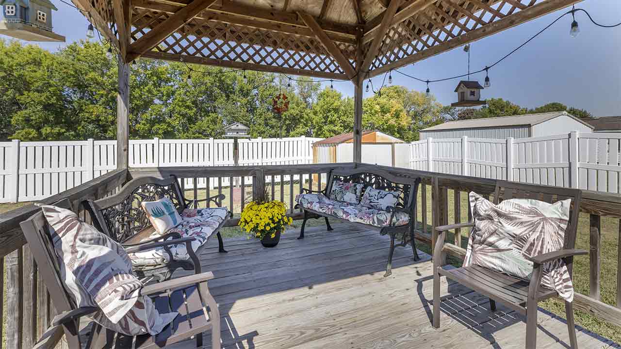 A wooden gazebo with a bench and chairs decorated with patterned cushions, a pot of yellow flowers in the center, string lights above, and a white fence and trees in the background.