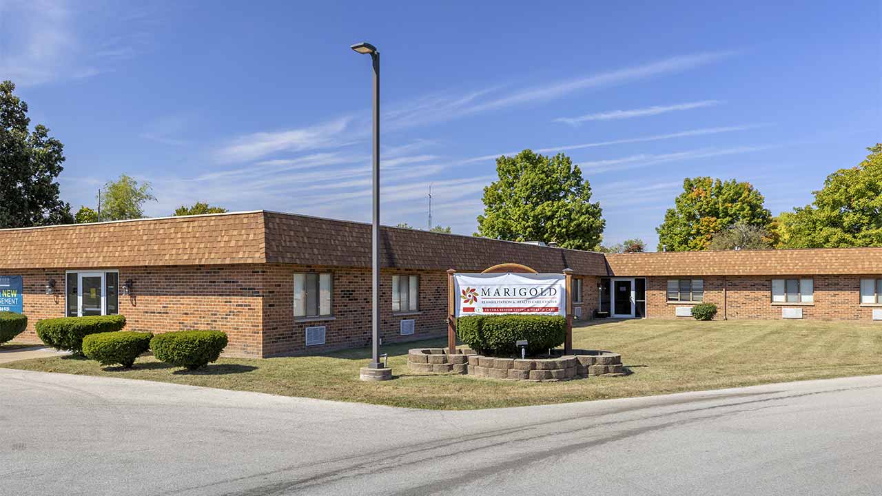 A one-story brick building with a brown shingle roof, surrounded by trimmed bushes and grass. A sign in front reads “Marigold.” Trees and a clear blue sky are in the background.