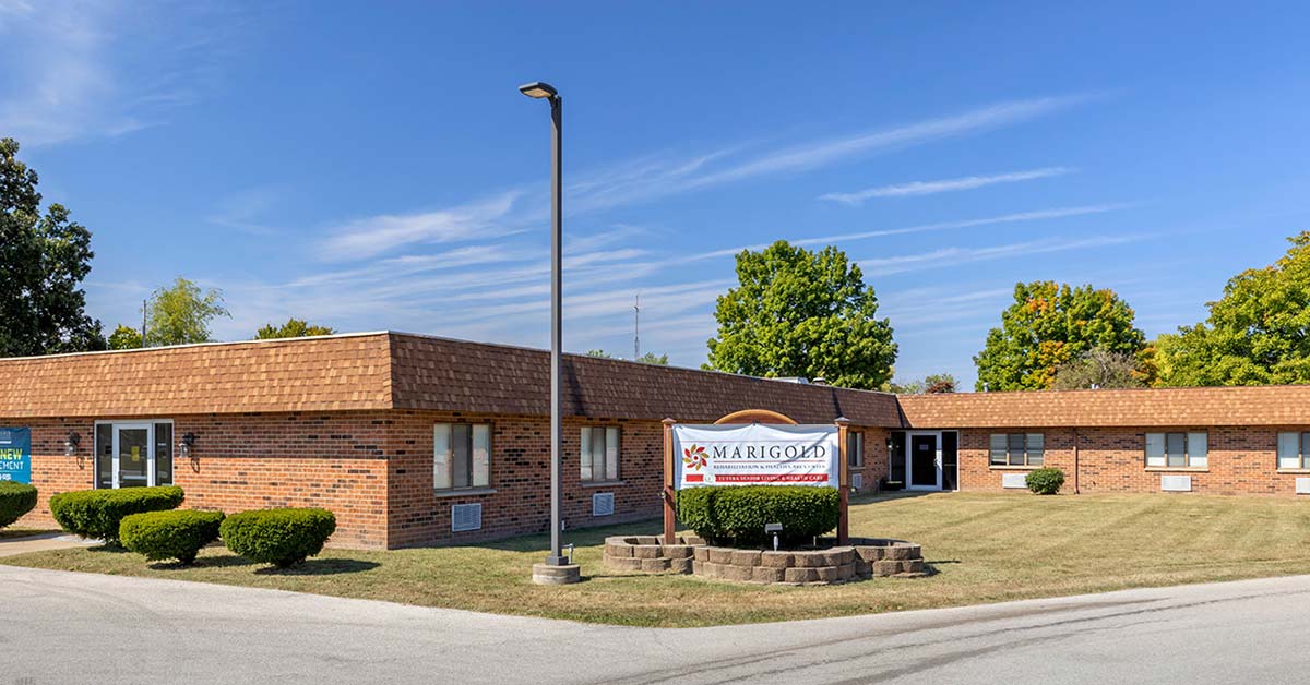 A one-story brick building with a shingled roof, surrounded by trimmed bushes and trees. A sign in front reads "Marigold." The sky is clear and blue.