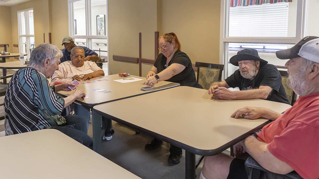 Five older adults and a younger woman sit around two tables playing cards in a brightly lit room with beige walls and large windows. The atmosphere appears relaxed and friendly.