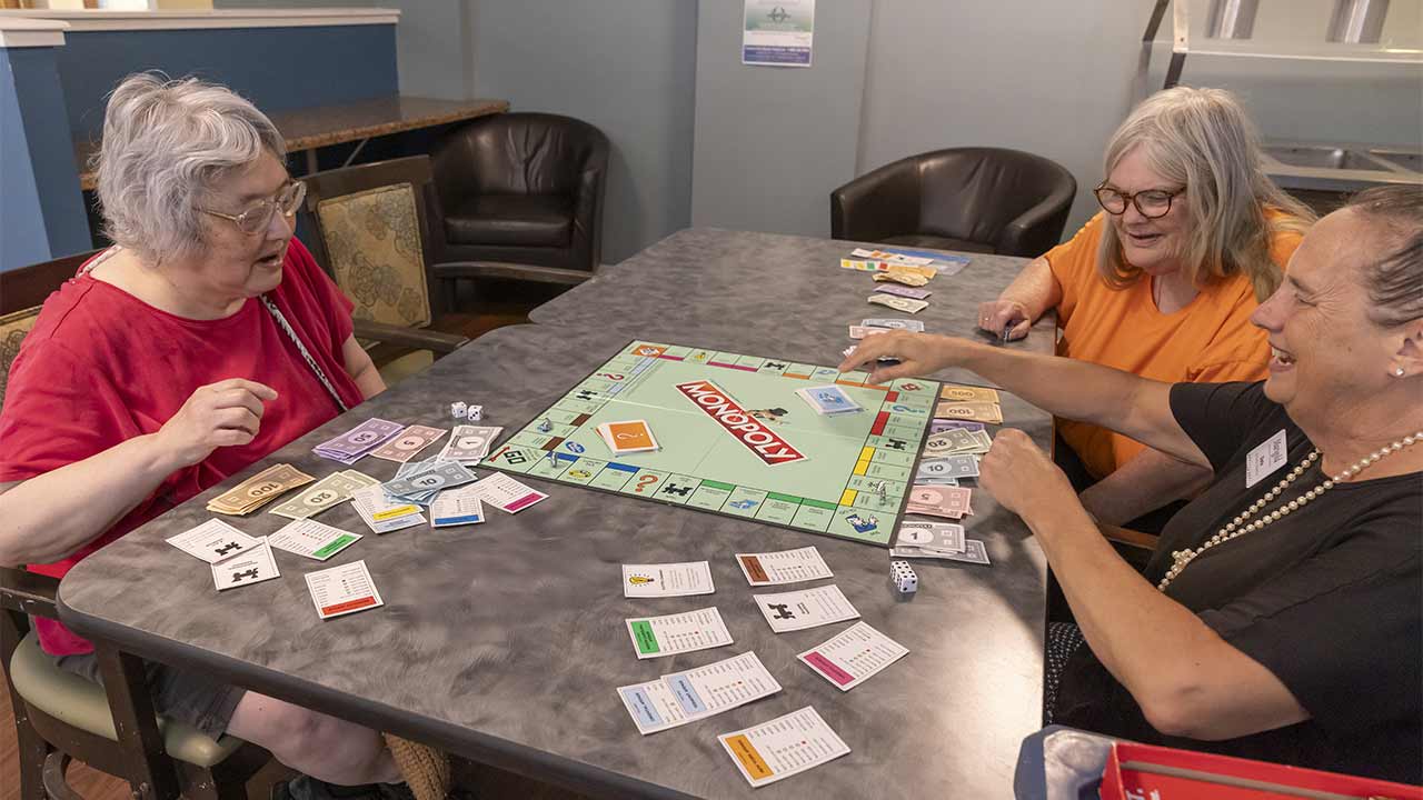 Three older women sit around a table, smiling and playing a game of Monopoly. Monopoly money, cards, and game pieces are spread out on the table. The women appear to be enjoying themselves in a comfortable indoor setting.