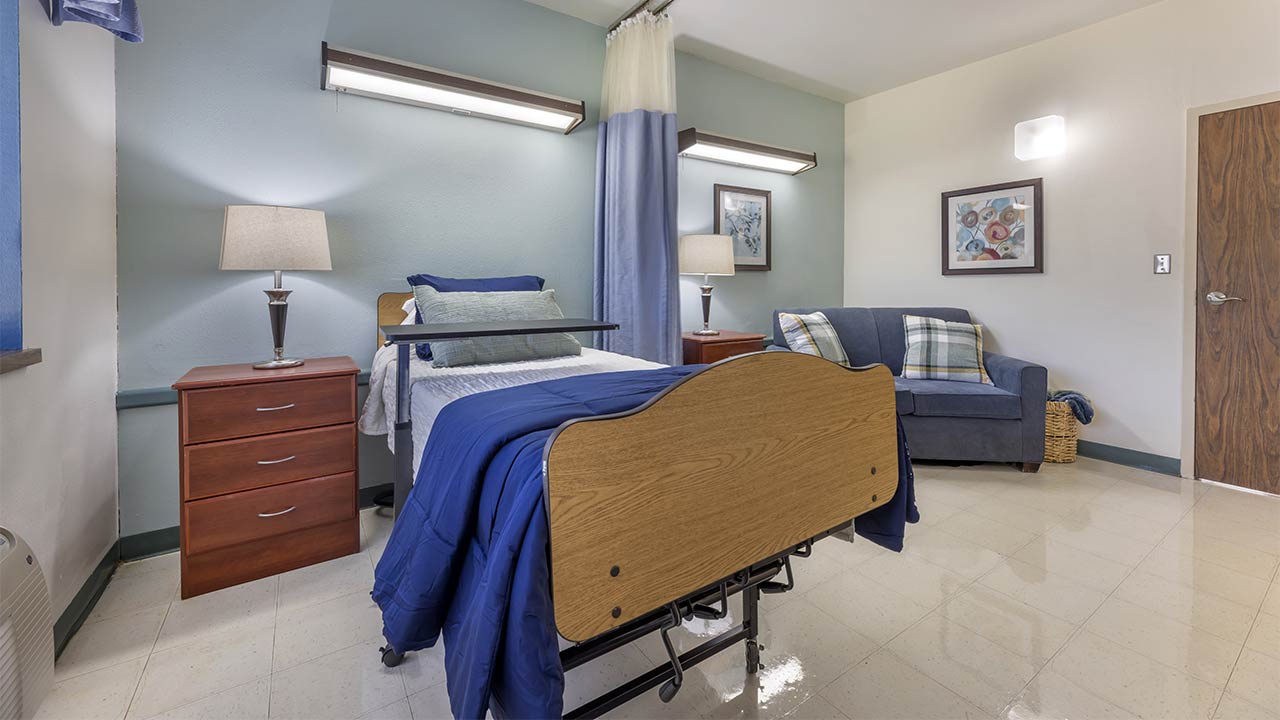 A clean, well-lit hospital or care facility room with a single bed, blue bedding, side tables with lamps, a blue couch with pillows, and framed art on the walls.