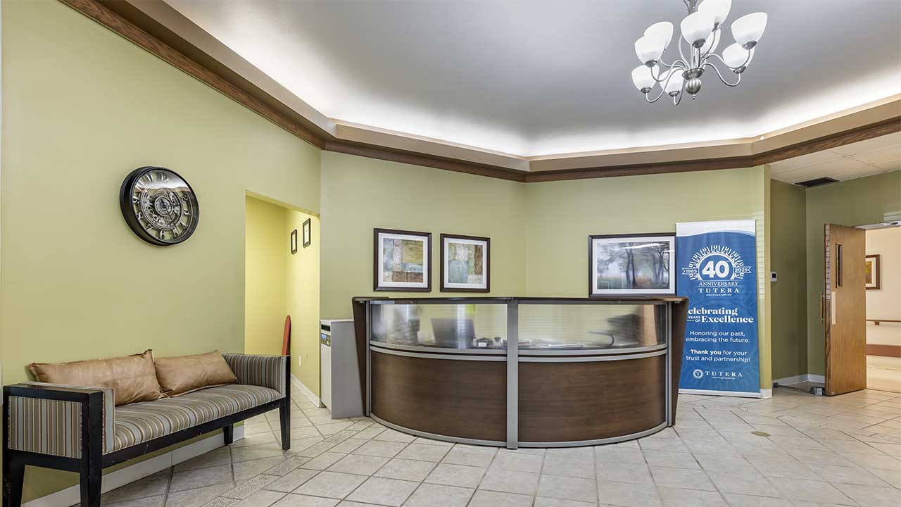 A reception area with a curved front desk, a couch with striped cushions, wall art, a clock, a banner celebrating a 40th anniversary, and a ceiling light fixture in a room with light green walls and tile flooring.