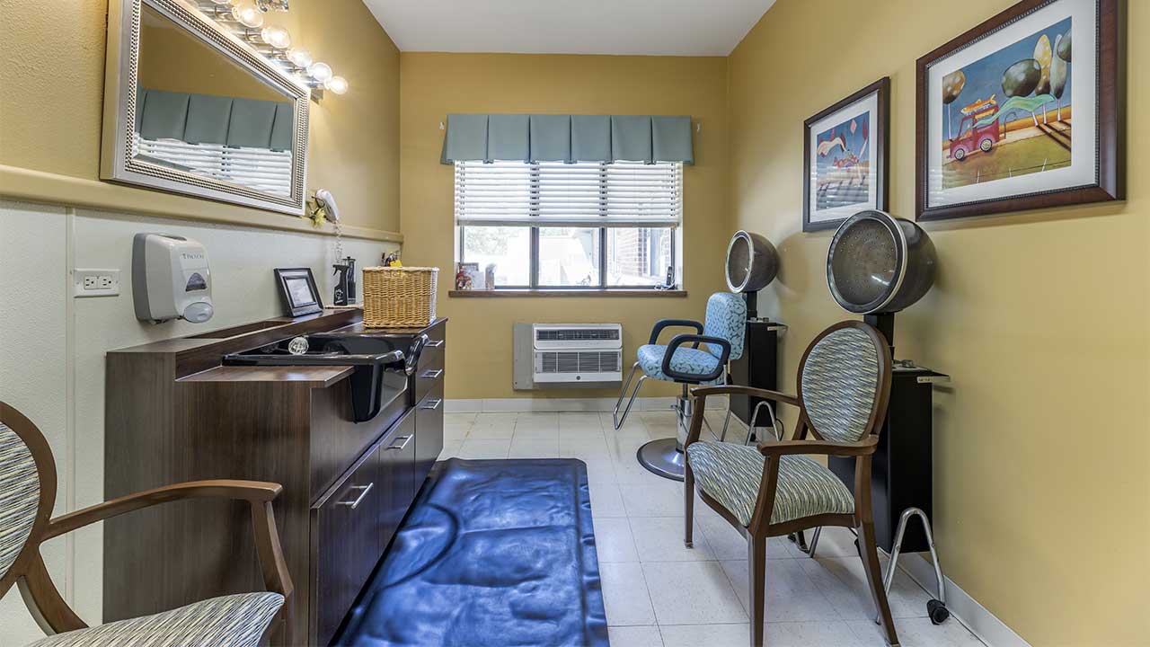 Small salon room with two hair dryer chairs, a vanity with a mirror, wooden cabinetry, and framed artwork on yellow walls. A window with a valance lets in natural light above an air conditioner.