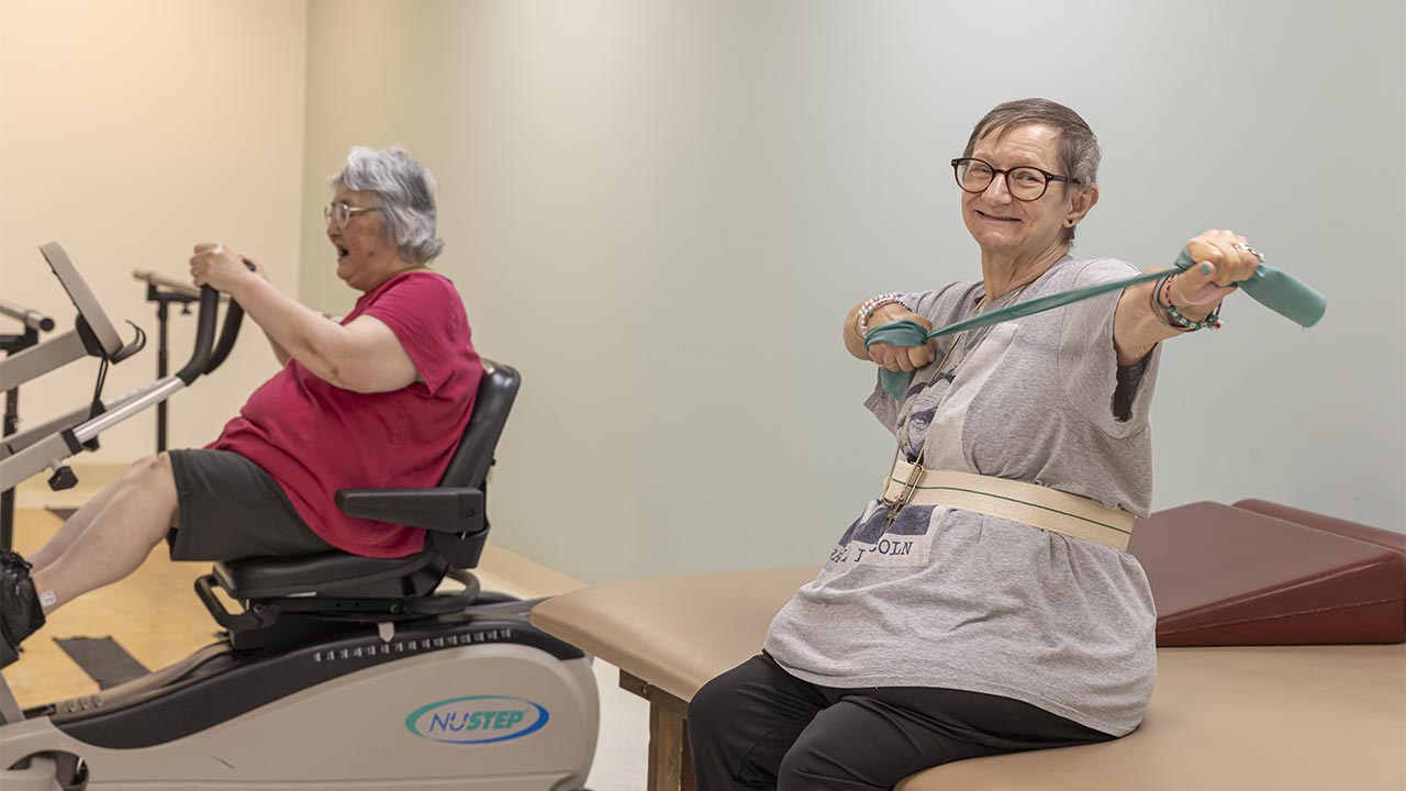 Two older adults exercise indoors; one uses a stationary bike, while the other, sitting on a mat table, smiles and stretches an exercise band sideways. Both appear engaged in physical therapy activities.