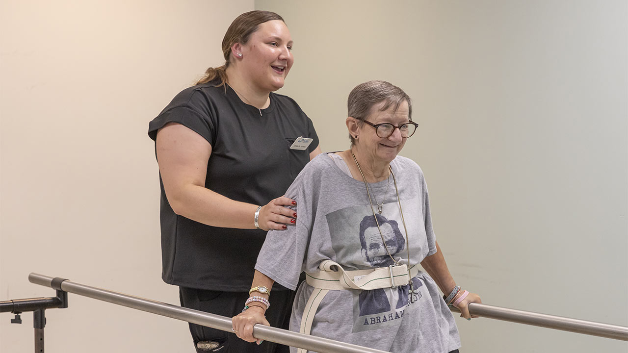 A smiling healthcare worker assists an older woman with glasses, who is walking between parallel bars during a physical therapy session. The woman wears a T-shirt with a printed image and a white gait belt for support.