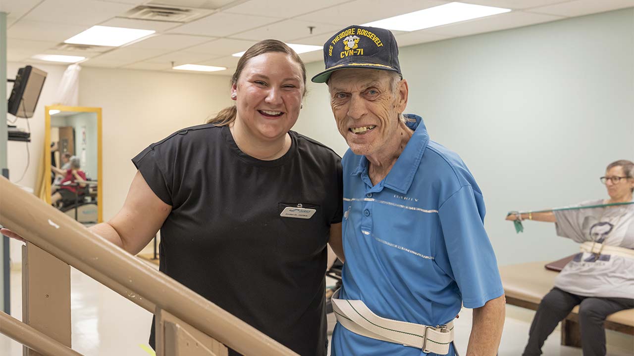 A smiling woman stands next to an elderly man in a navy cap and blue shirt at a rehabilitation center. Both look happy; the man has a gait belt on, and another person exercises with resistance bands in the background.