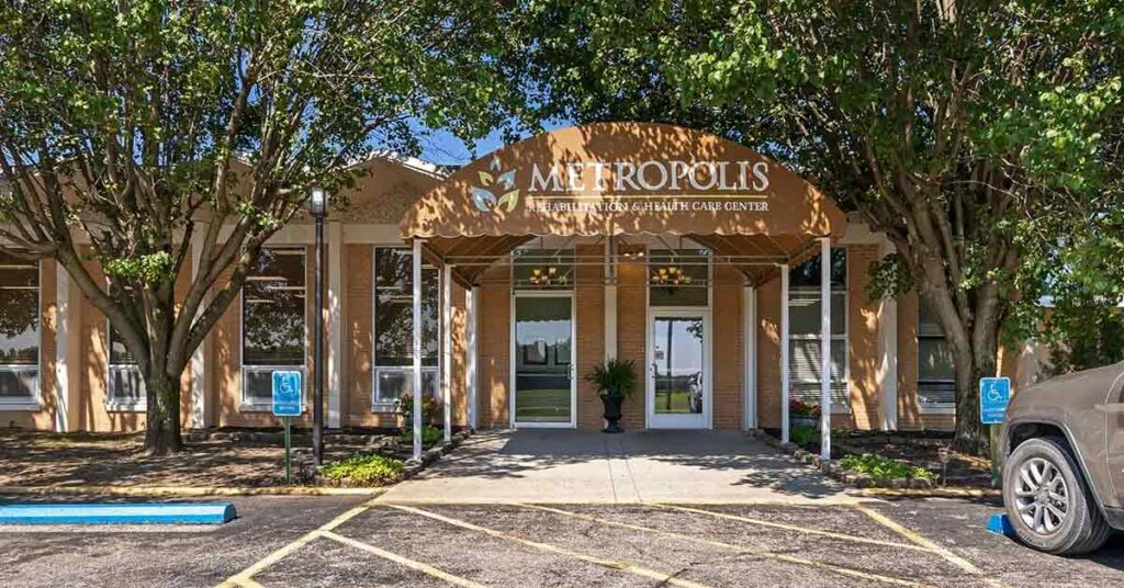 The entrance of Metropolis Rehabilitation & Health Care Center features a brown awning, glass double doors, large windows, two handicap parking spaces, and trees shading the health care center.