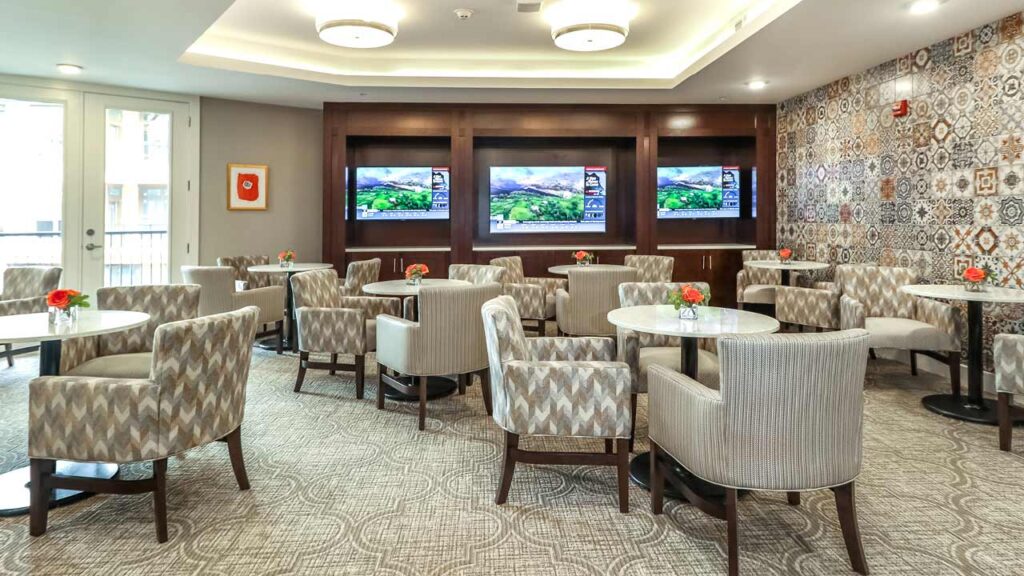 A modern, well-lit lounge area designed for senior living, with numerous round tables and cushioned chairs arranged neatly. Three flat-screen TVs are mounted on a wooden wall displaying scenic images. The room features patterned wallpaper on one side and small red floral arrangements on the tables.