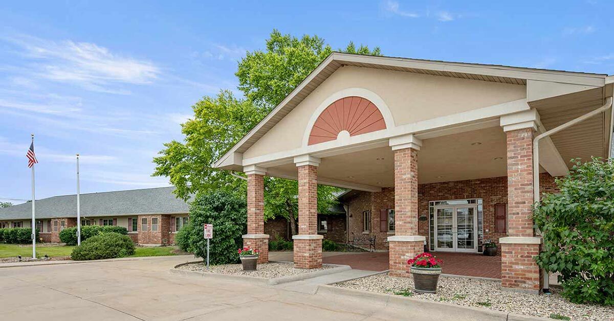 Single-story brick building with a covered entrance supported by columns, potted flowers near the entrance, green trees, a U.S. flag, and a clear blue sky in the background.
