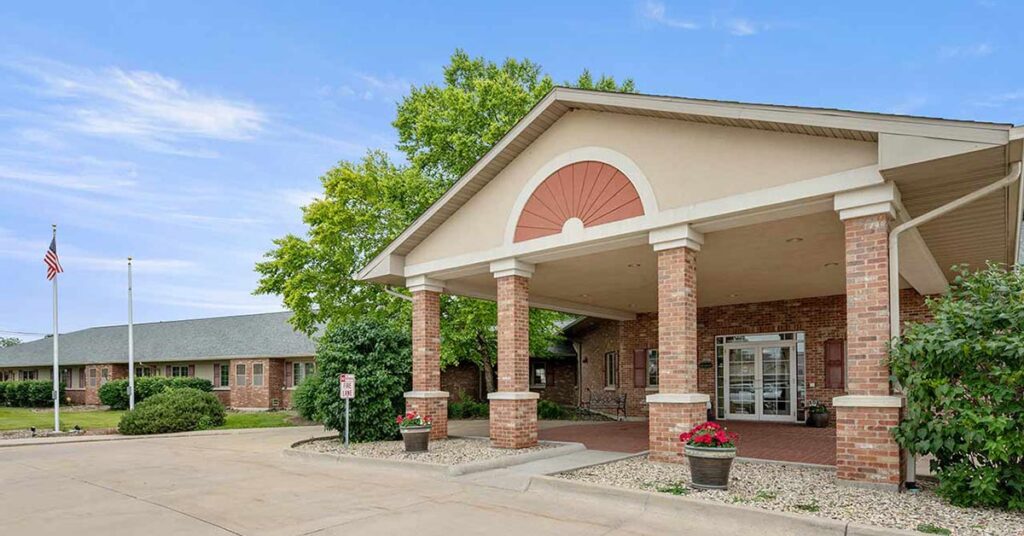 Single-story brick building with a covered entrance supported by columns, potted flowers near the entrance, green trees, a U.S. flag, and a clear blue sky in the background.
