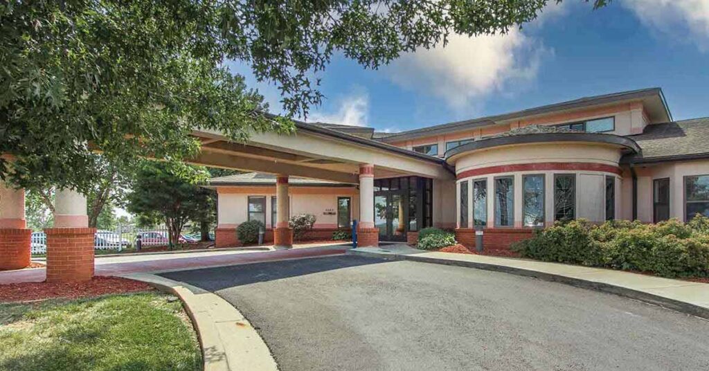 Single-story Rehabilitation & Health Care Center with a covered entrance and circular windows, surrounded by bushes and trees. A driveway leads to the entrance under a large awning beneath a partly cloudy sky.