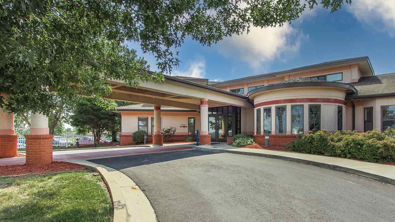 A modern Rehabilitation & Health Care Center with large windows and a covered entrance driveway supported by red columns, surrounded by green trees, shrubs, and a well-maintained lawn under a partly cloudy sky.