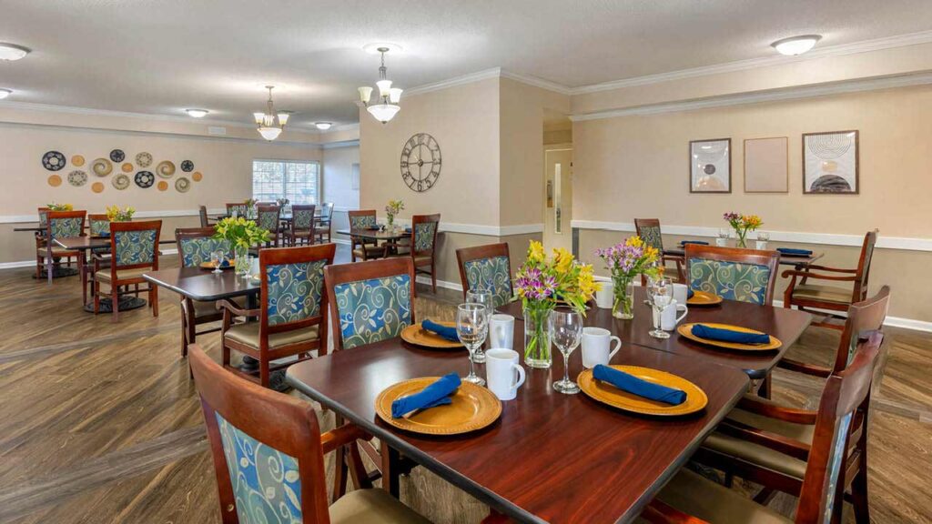 A spacious, well-lit dining room with wooden tables set with gold chargers, white mugs, glasses, and navy napkins. The chairs have blue patterned cushions and fresh flowers decorate each table. Artwork and clocks adorn the walls.