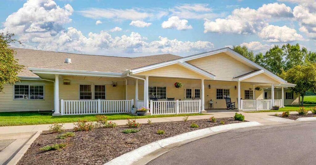 A single-story, light yellow building with a white porch railing, columns, and a gabled roof, surrounded by landscaping and a curved driveway under a partly cloudy sky.