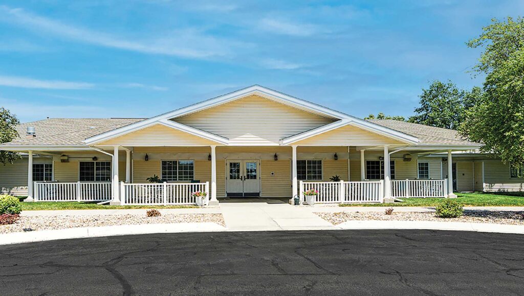 A single-story beige building with a gabled roof, featuring a spacious front porch lined with white railings and columns. The building is set back from the road, with a wide paved driveway in front and landscaped areas with green grass and young trees.