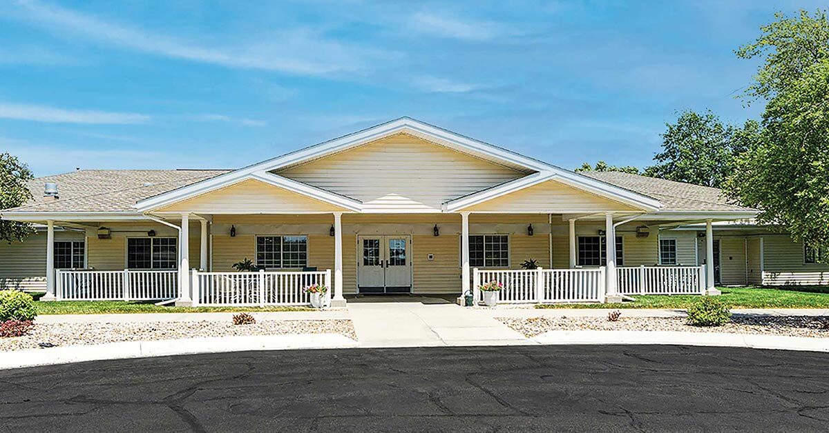 A single-story yellow house with a gabled roof and a spacious front porch. The porch is supported by white pillars, and the house is surrounded by a neatly maintained lawn and shrubs. A paved driveway leads to the entrance.